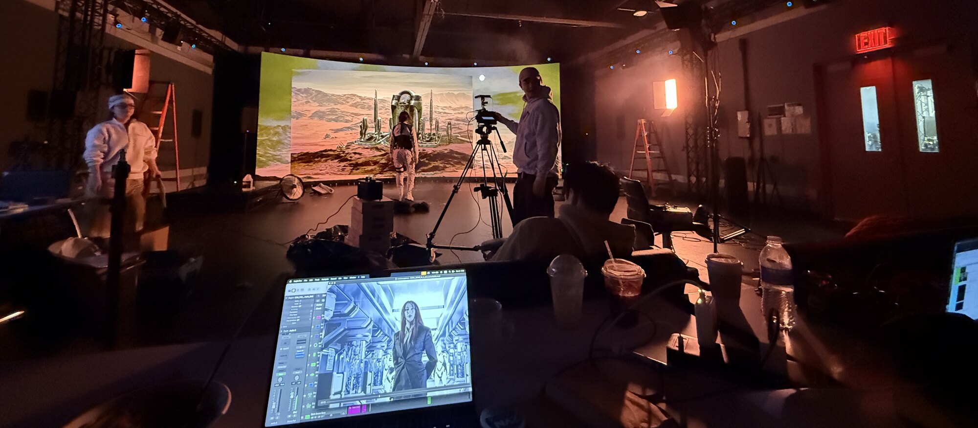 A film set: a projected desert backdrop, a camera on a tripod, and an editing timeline open on a laptop in the foreground