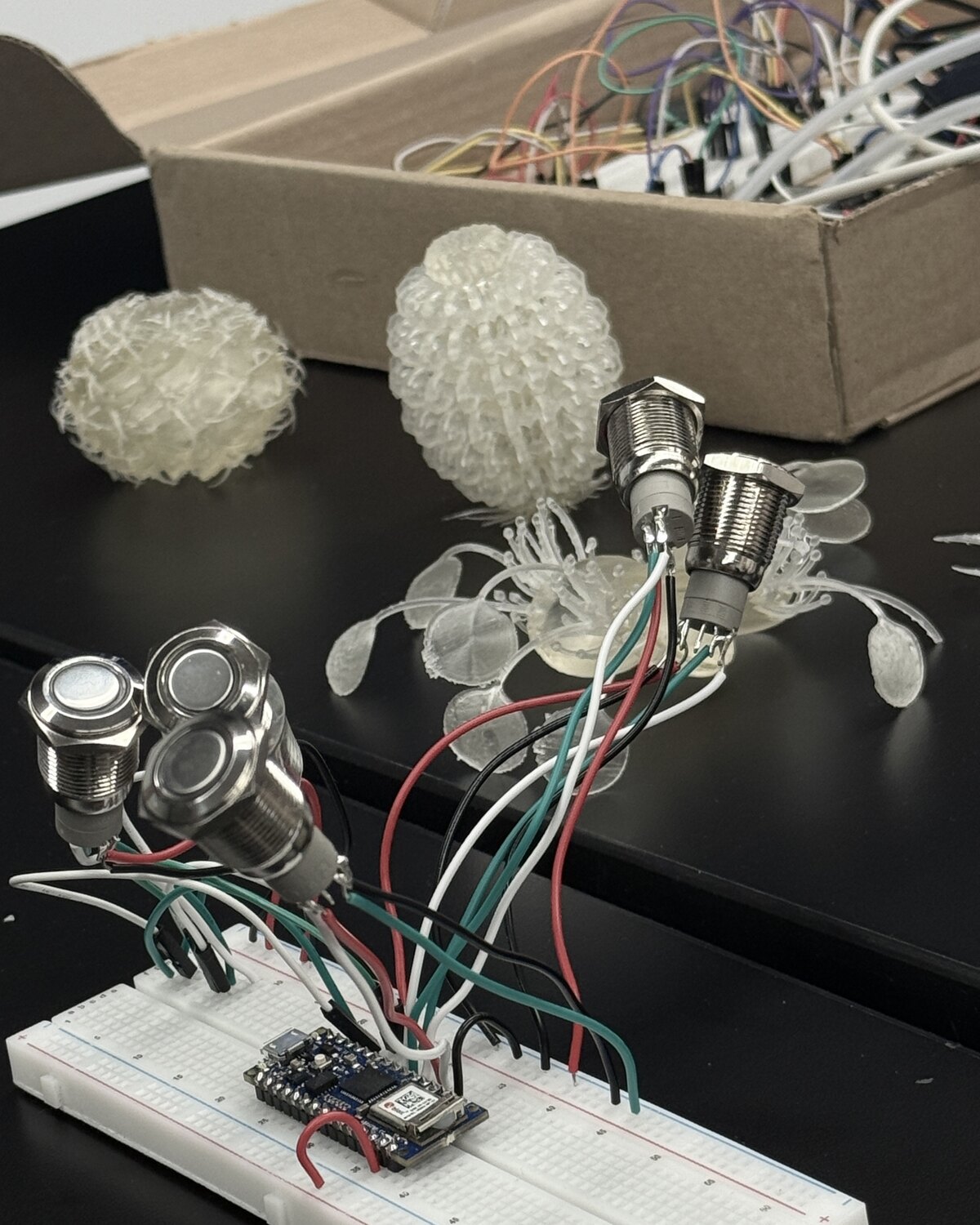 A workbench with 3D-printed translucent forms, arcade buttons and a wired breadboard