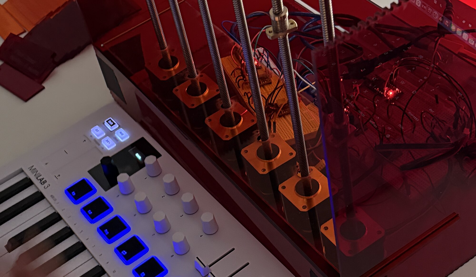 A studio bench: a red acrylic machine driven by stepper motors and threaded rods, wired to a microcontroller, with a hand playing a MIDI keyboard beside it